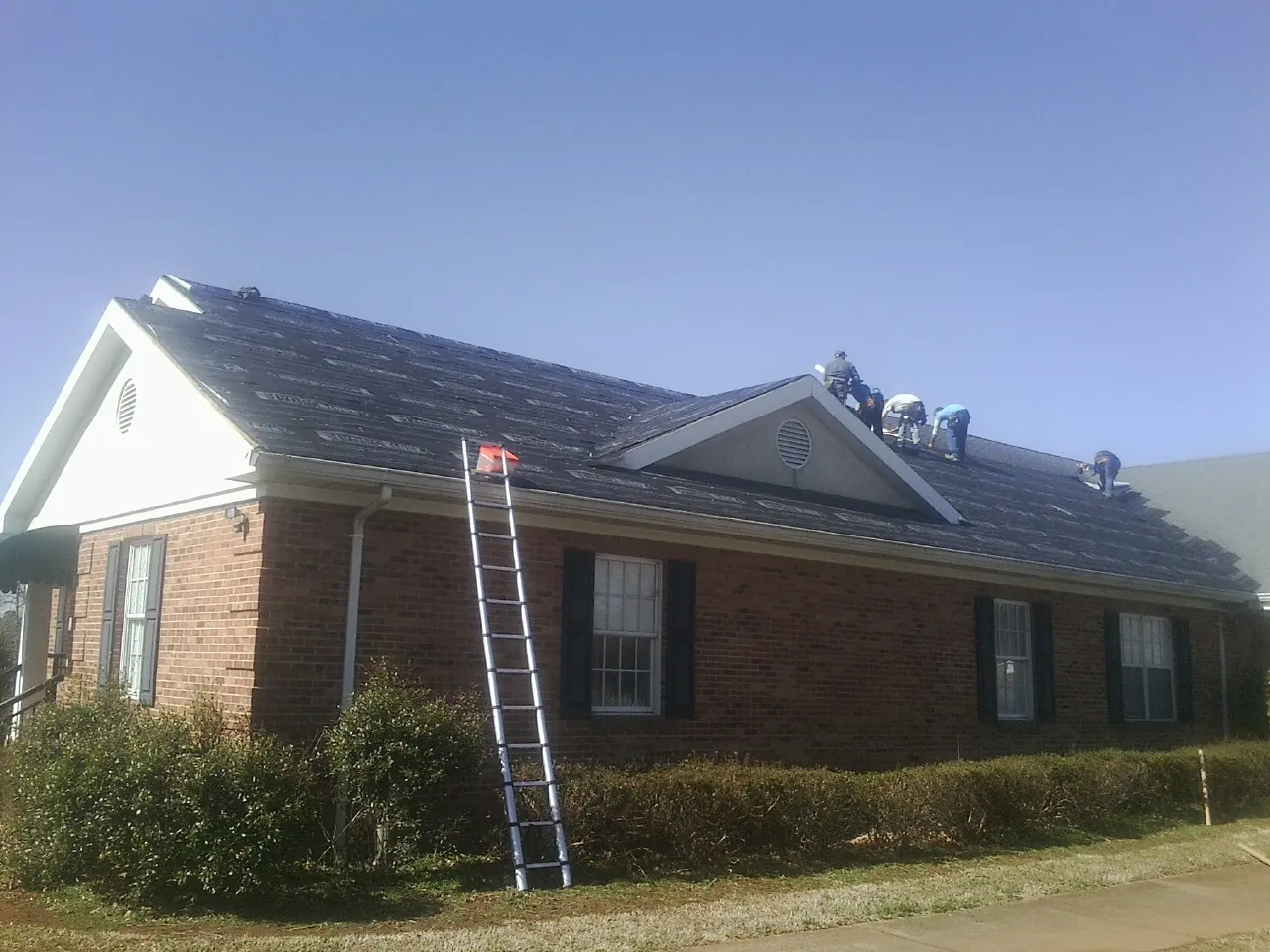 Crew tearing shingles off a brick ranch roof with a ladder set — Metal Roof Repair work by our crew in Dumont